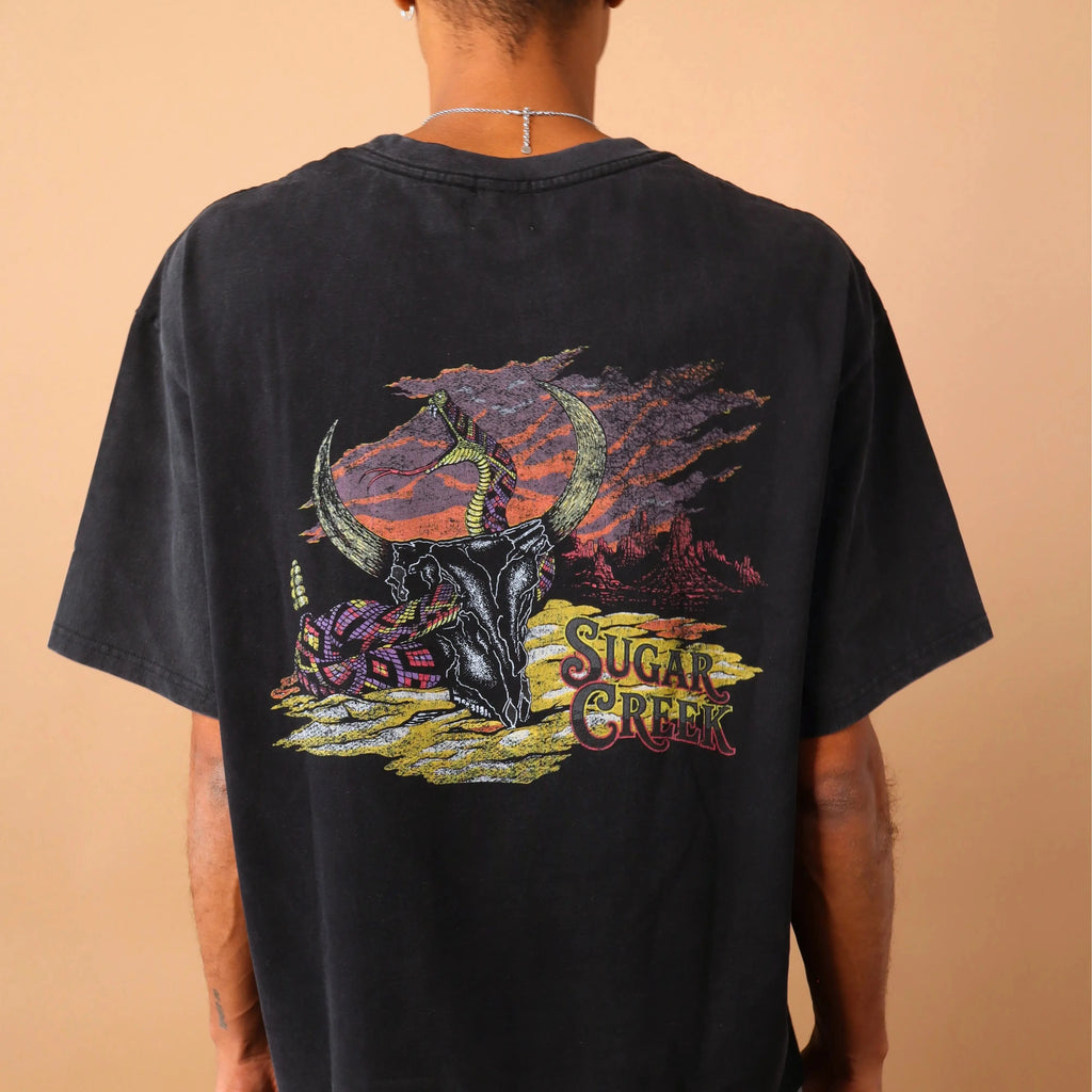 Person wearing a black t-shirt with a colorful graphic design on the back, standing against a beige wall.