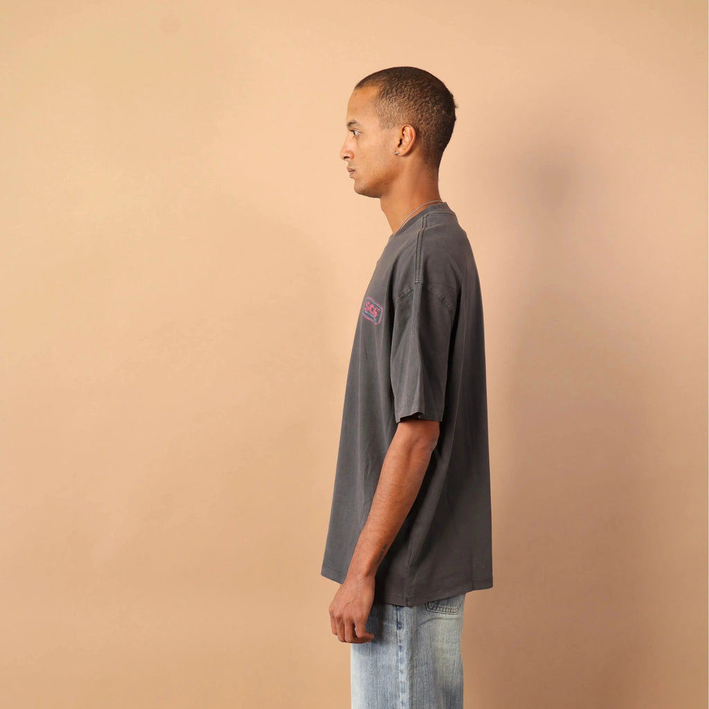 Man wearing a gray t-shirt and light blue jeans standing against a beige wall.
