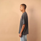 Man wearing a gray t-shirt and light blue jeans standing against a beige wall.