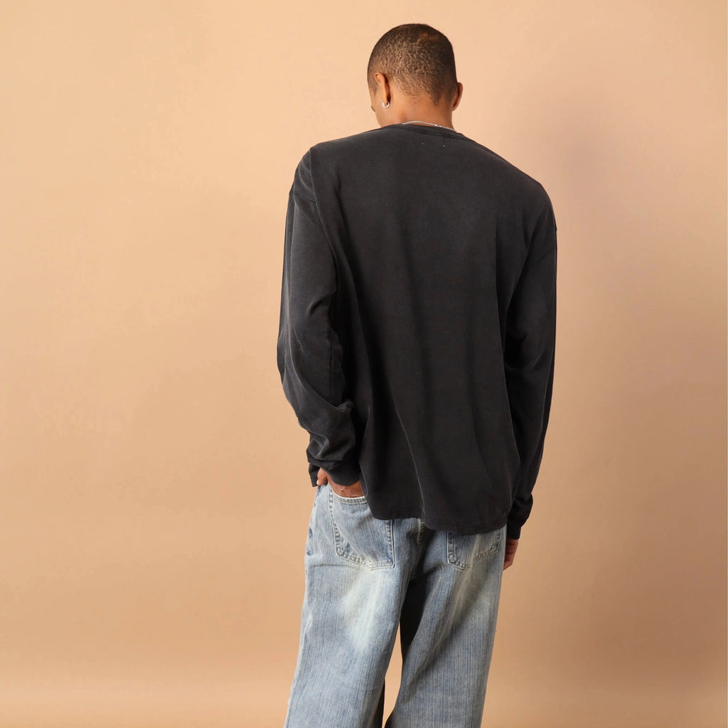 Person wearing a black long-sleeve shirt and light blue jeans against a beige wall.