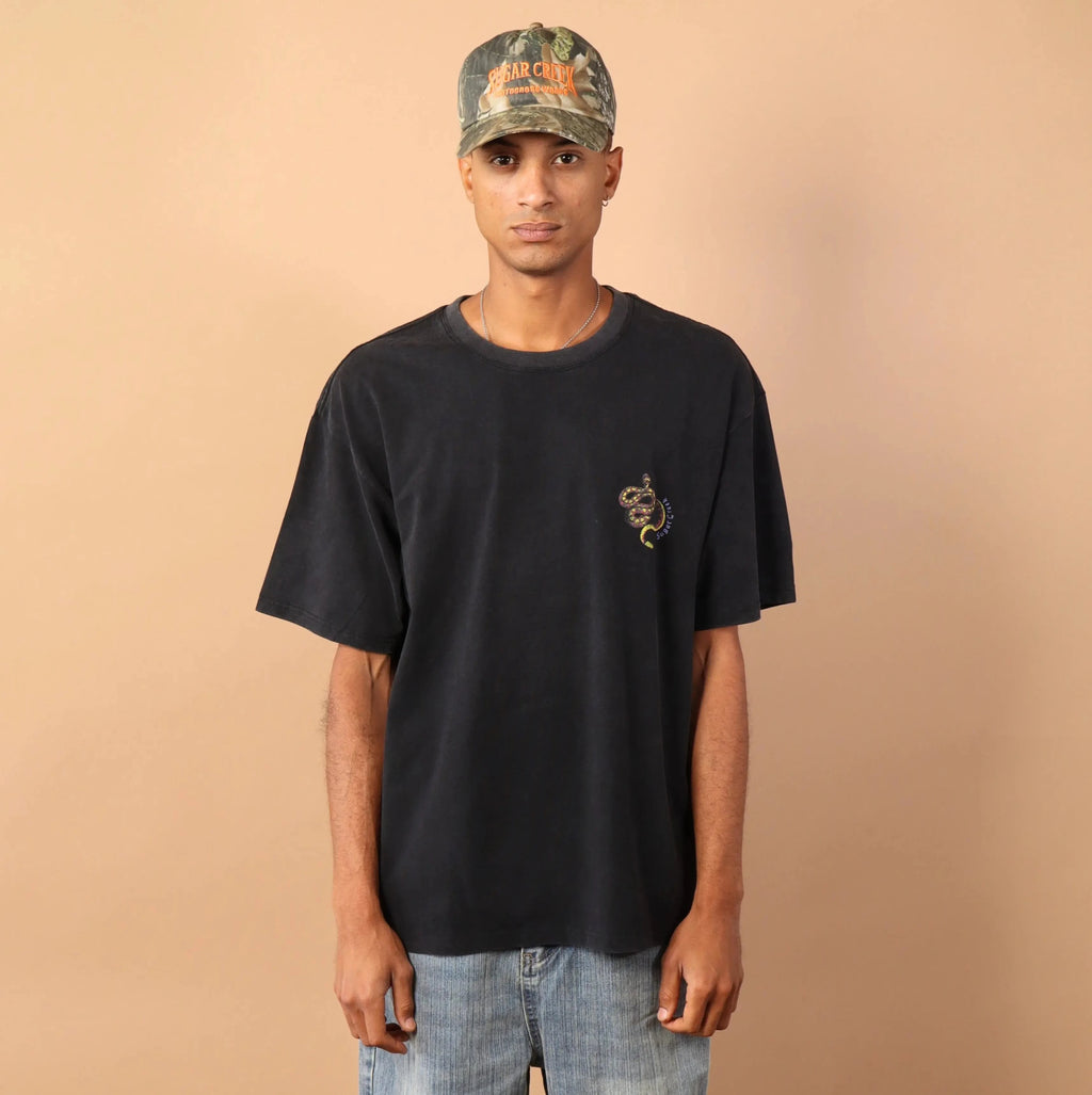Person wearing a black t-shirt, light blue jeans, and a camouflage cap against a beige background