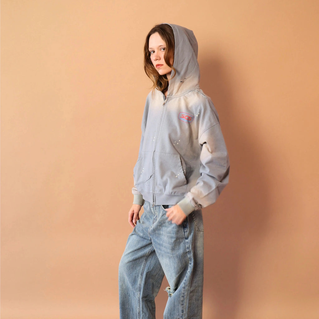Person wearing a light gray hoodie and blue jeans against a beige wall.