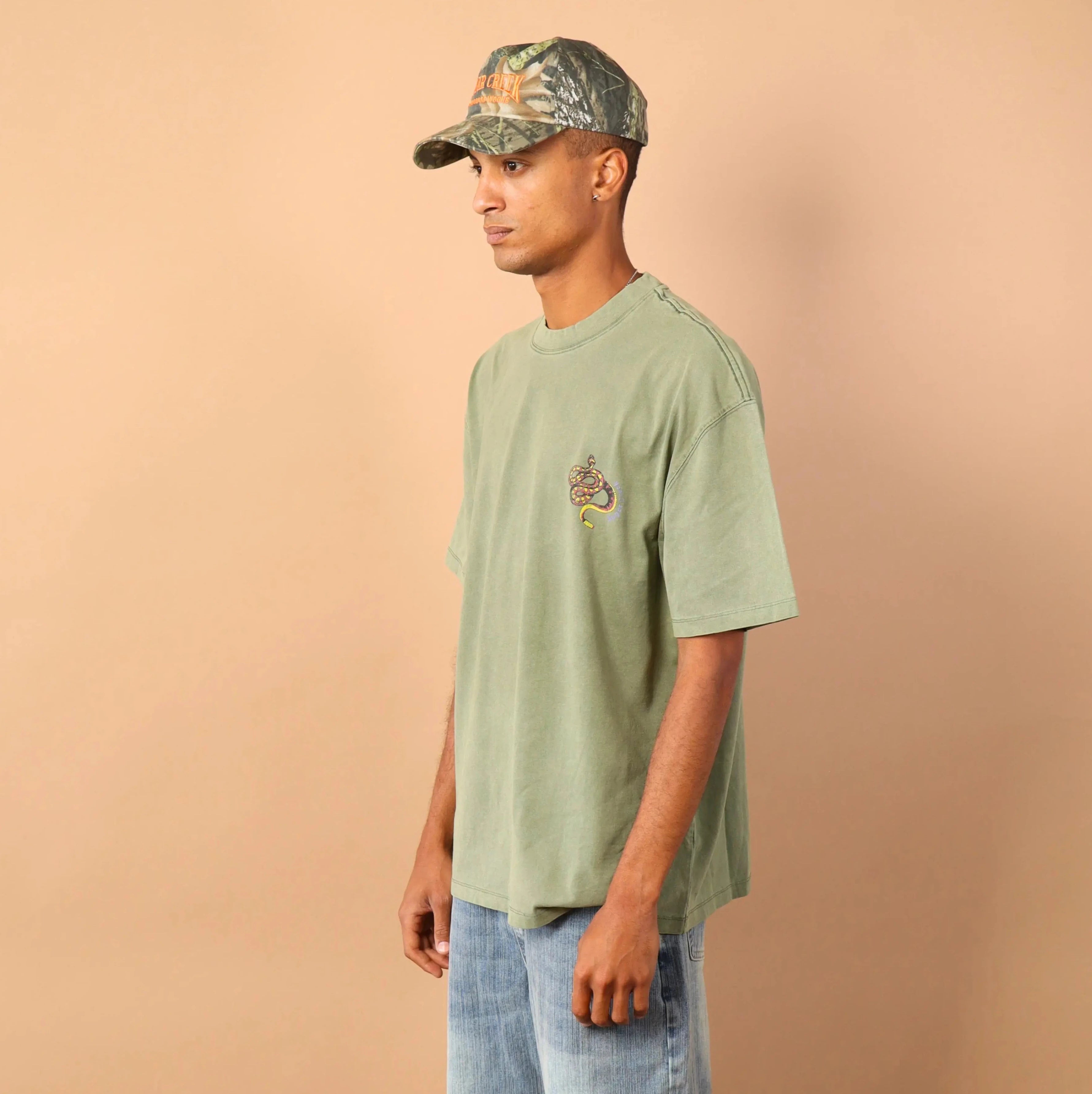 Person wearing a green t-shirt and blue jeans standing against a beige wall.