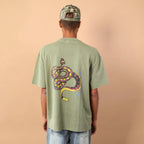 Person wearing a green t-shirt with a snake design on the back, standing against a beige wall.