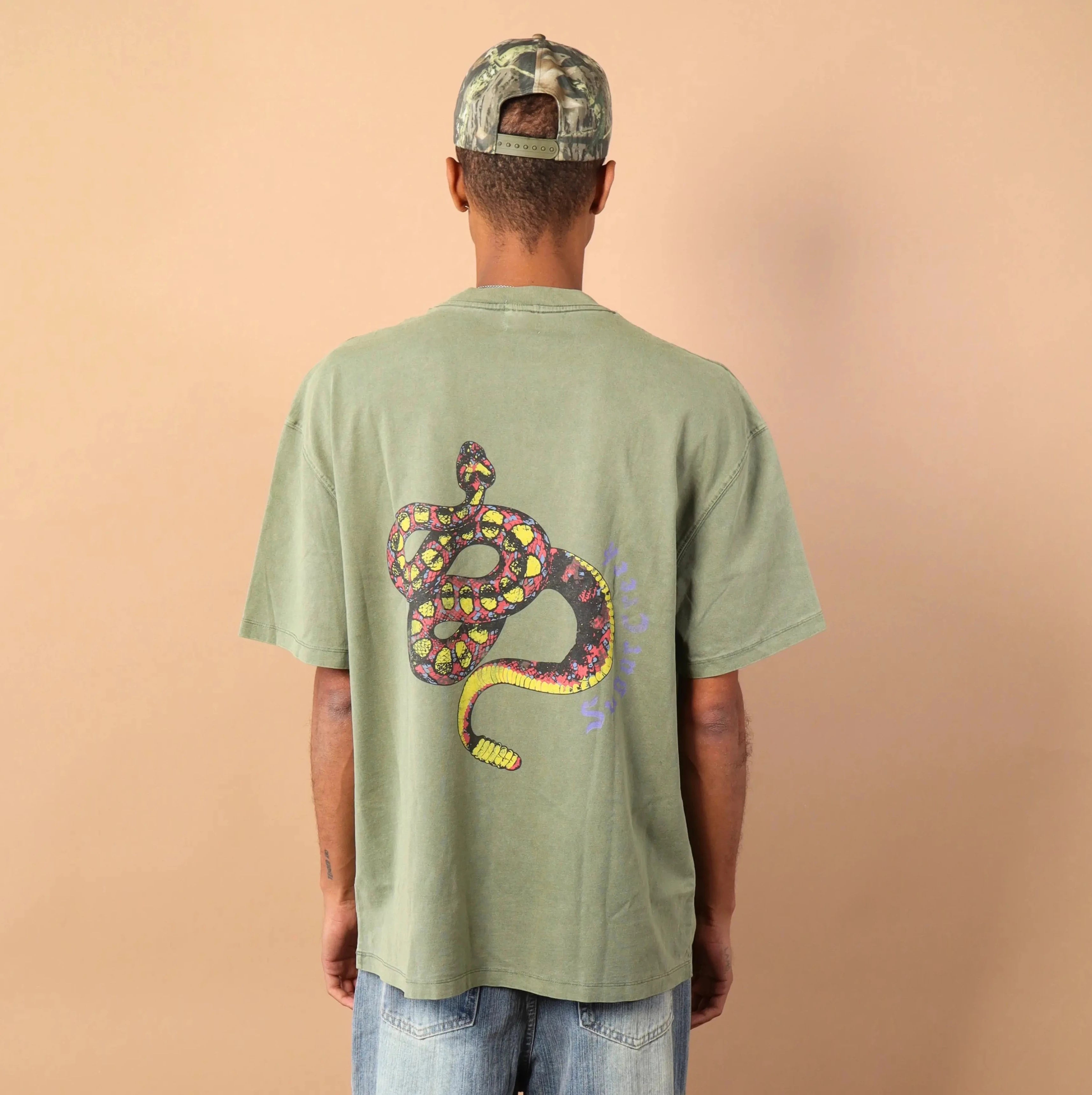 Person wearing a green t-shirt with a snake design on the back, standing against a beige wall.