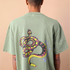 Person wearing a green t-shirt with a colorful snake design on the back, standing against a beige background.
