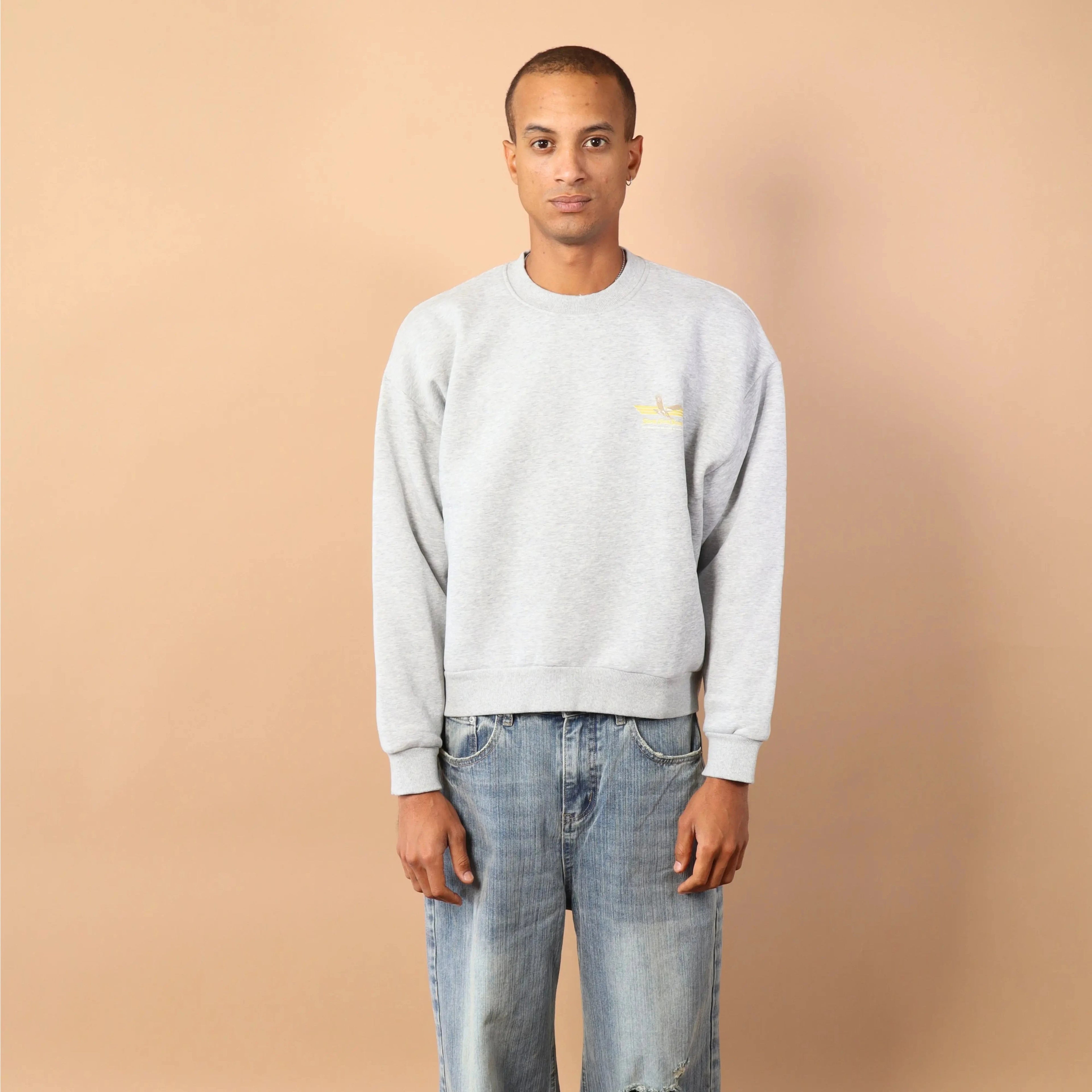 Man wearing a light gray sweatshirt and blue jeans standing against a beige wall.