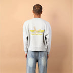 Person wearing a light gray sweatshirt with a logo on the back against a beige wall.
