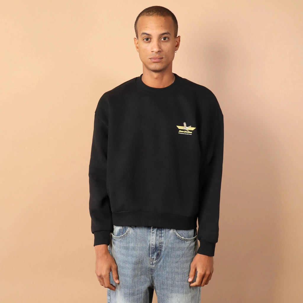 Man wearing a black sweater with a logo and light blue jeans against a beige background