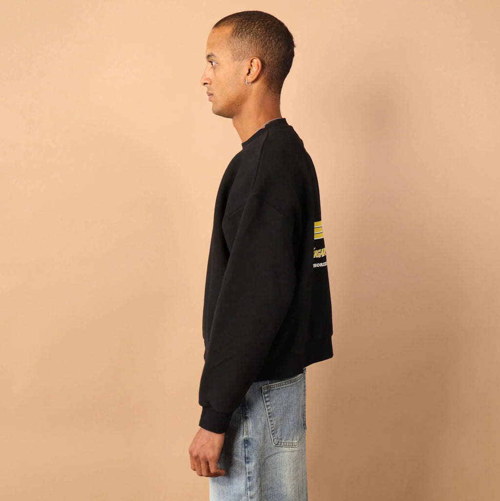 Person wearing a black sweatshirt with a gold stripe and light blue jeans against a beige wall.