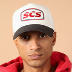 Person wearing a white cap with 'SCS' logo and red hoodie on beige background