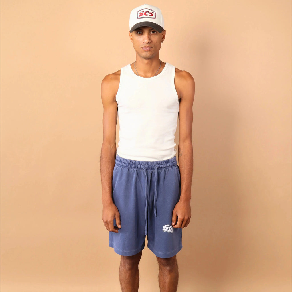 Person wearing a white tank top, blue shorts, and a cap against a beige background