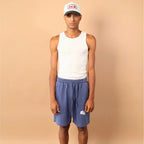 Person wearing a white tank top, blue shorts, and a cap against a beige background