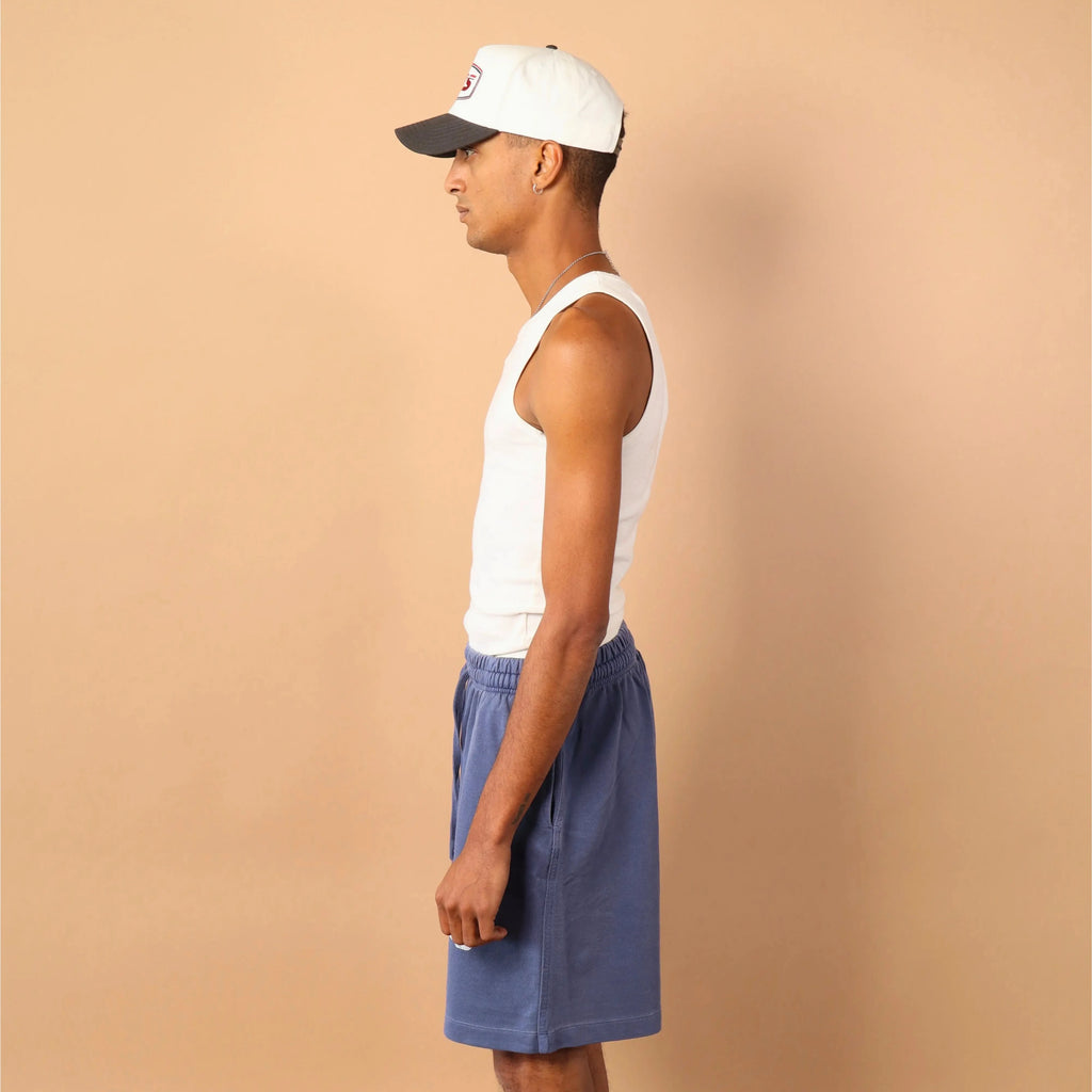 Man wearing a white tank top, blue shorts, and a cap against a beige background
