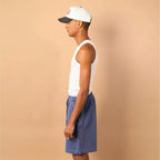 Man wearing a white tank top, blue shorts, and a cap against a beige background