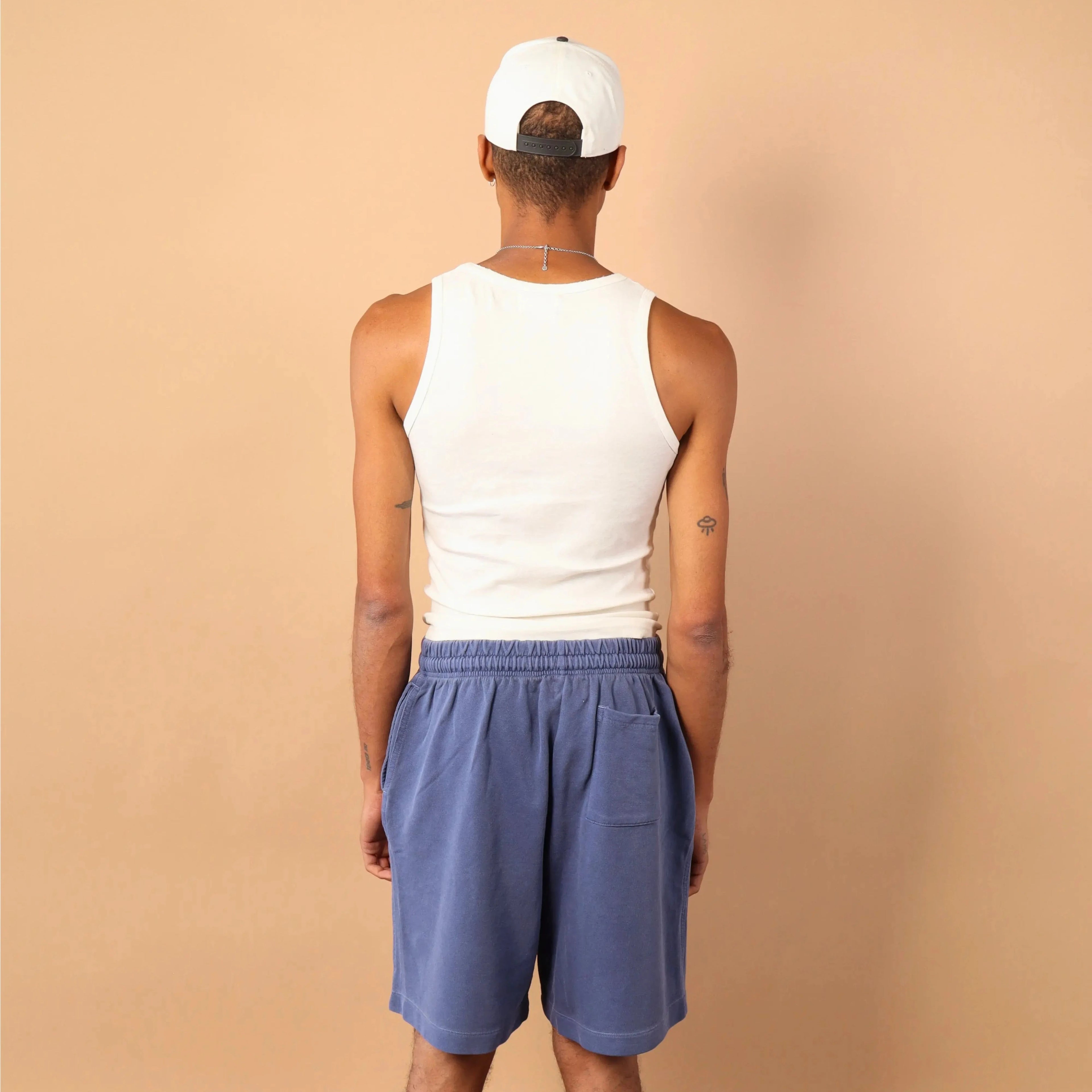 Person wearing a white tank top, blue shorts, and a white cap on a beige background