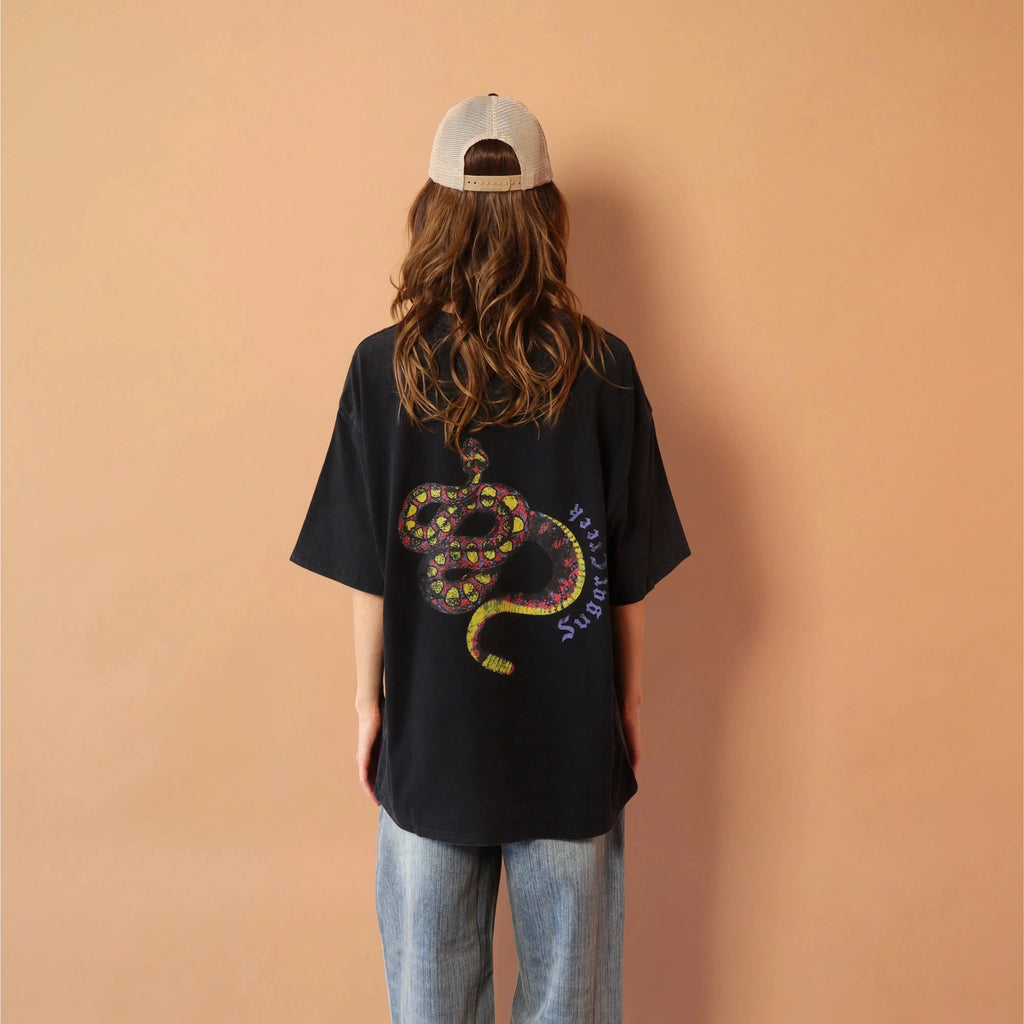 Person wearing a black t-shirt with a snake design, standing against a peach wall.