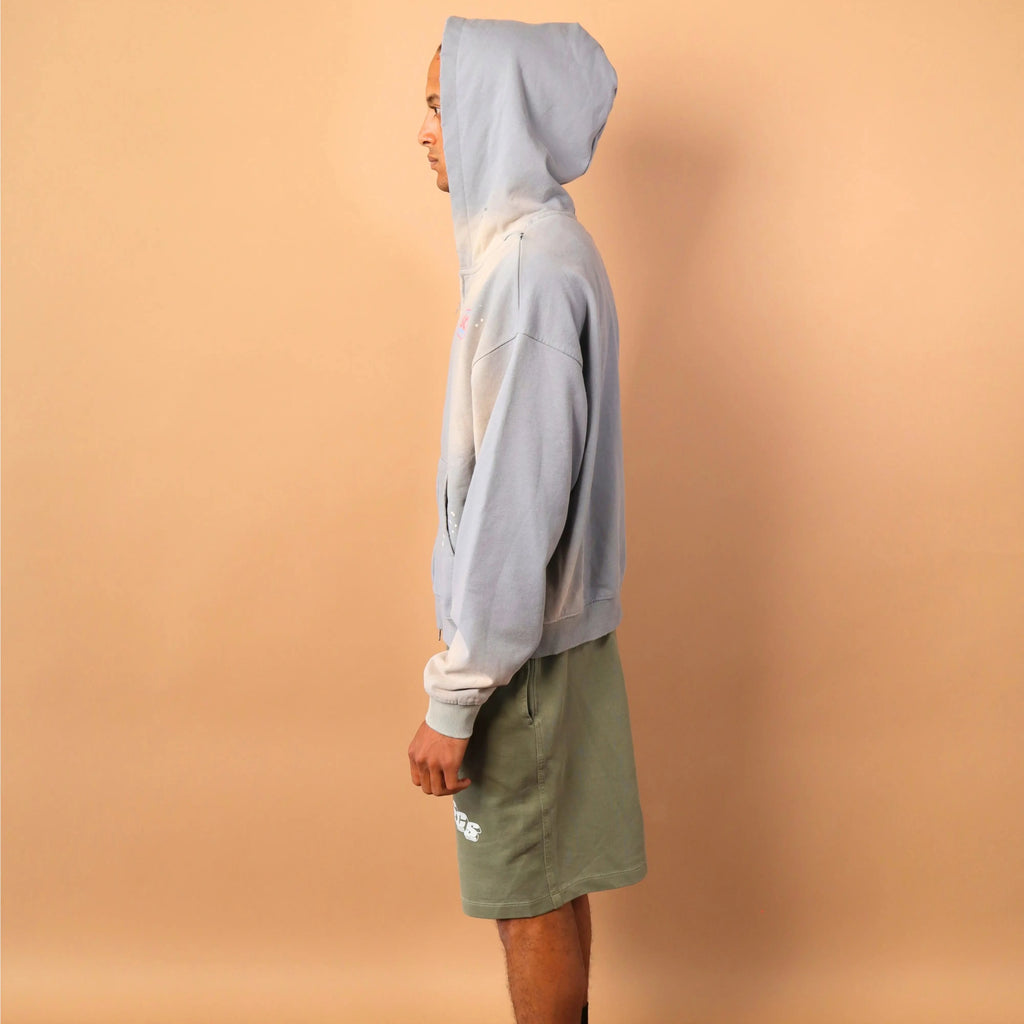 Person wearing a light gray hoodie and green shorts standing against a beige wall.