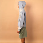 Person wearing a light gray hoodie and green shorts standing against a beige wall.