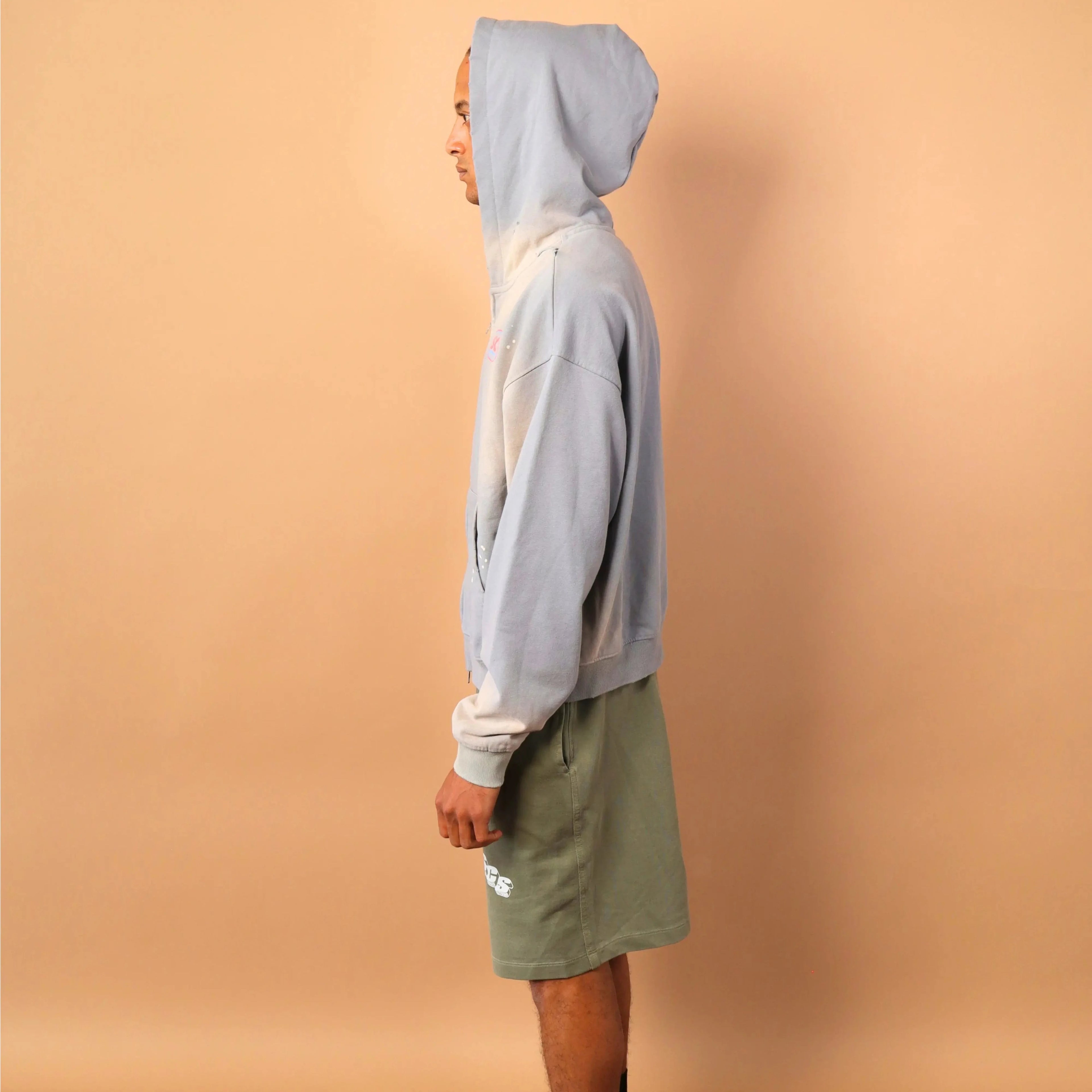 Person wearing a light gray hoodie and green shorts standing against a beige wall.