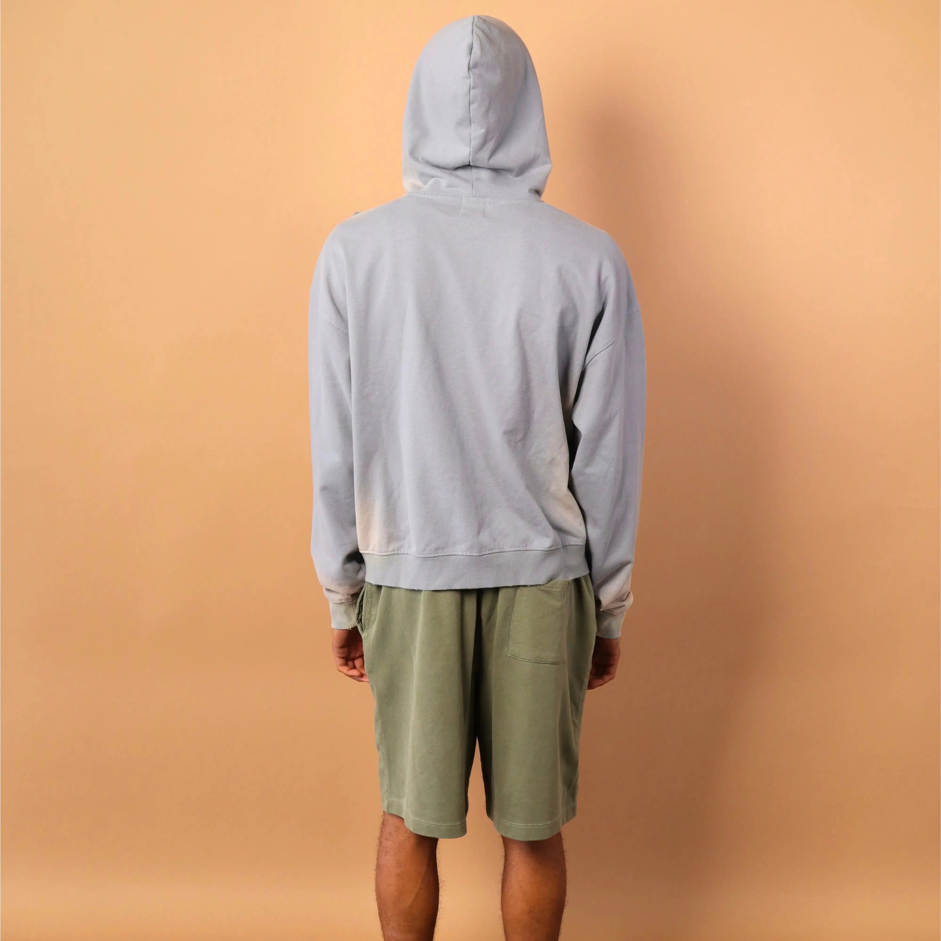 Person wearing a gray hoodie and green shorts standing against a beige wall.