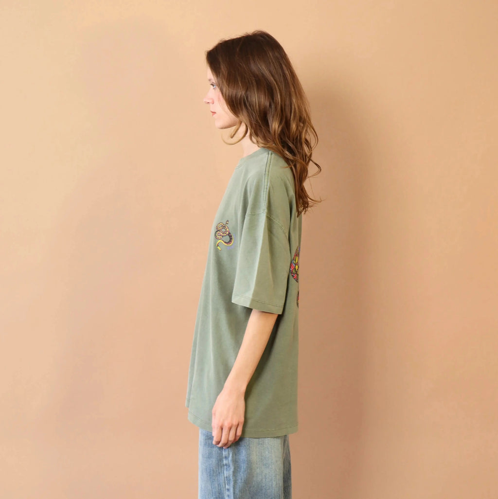 Person wearing a green oversized t-shirt and light blue jeans against a beige wall.