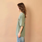 Person wearing a green oversized t-shirt and light blue jeans against a beige wall.