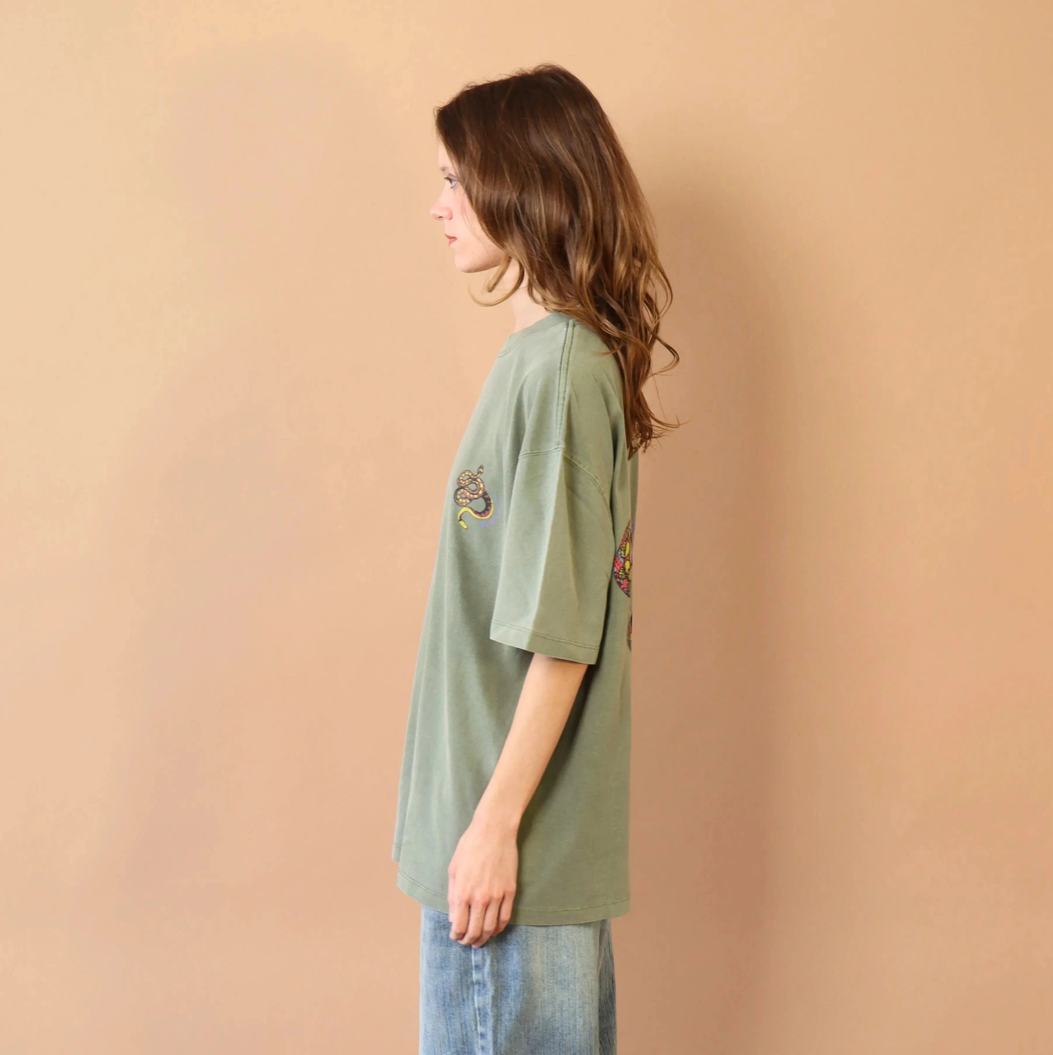 Person wearing a green oversized t-shirt and light blue jeans against a beige wall.