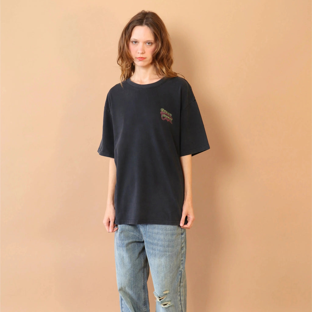 Person wearing a black t-shirt and blue jeans against a beige wall.
