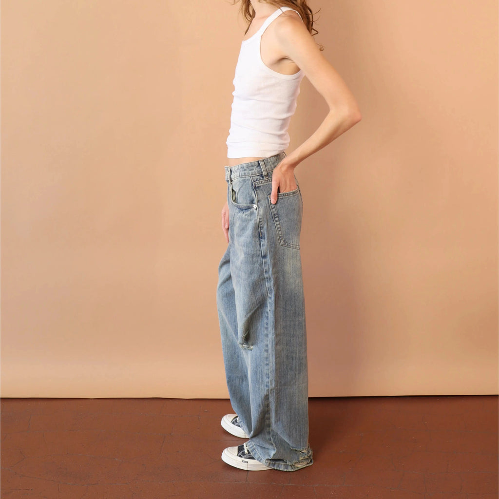 Woman wearing a white tank top and blue jeans standing against a beige wall.