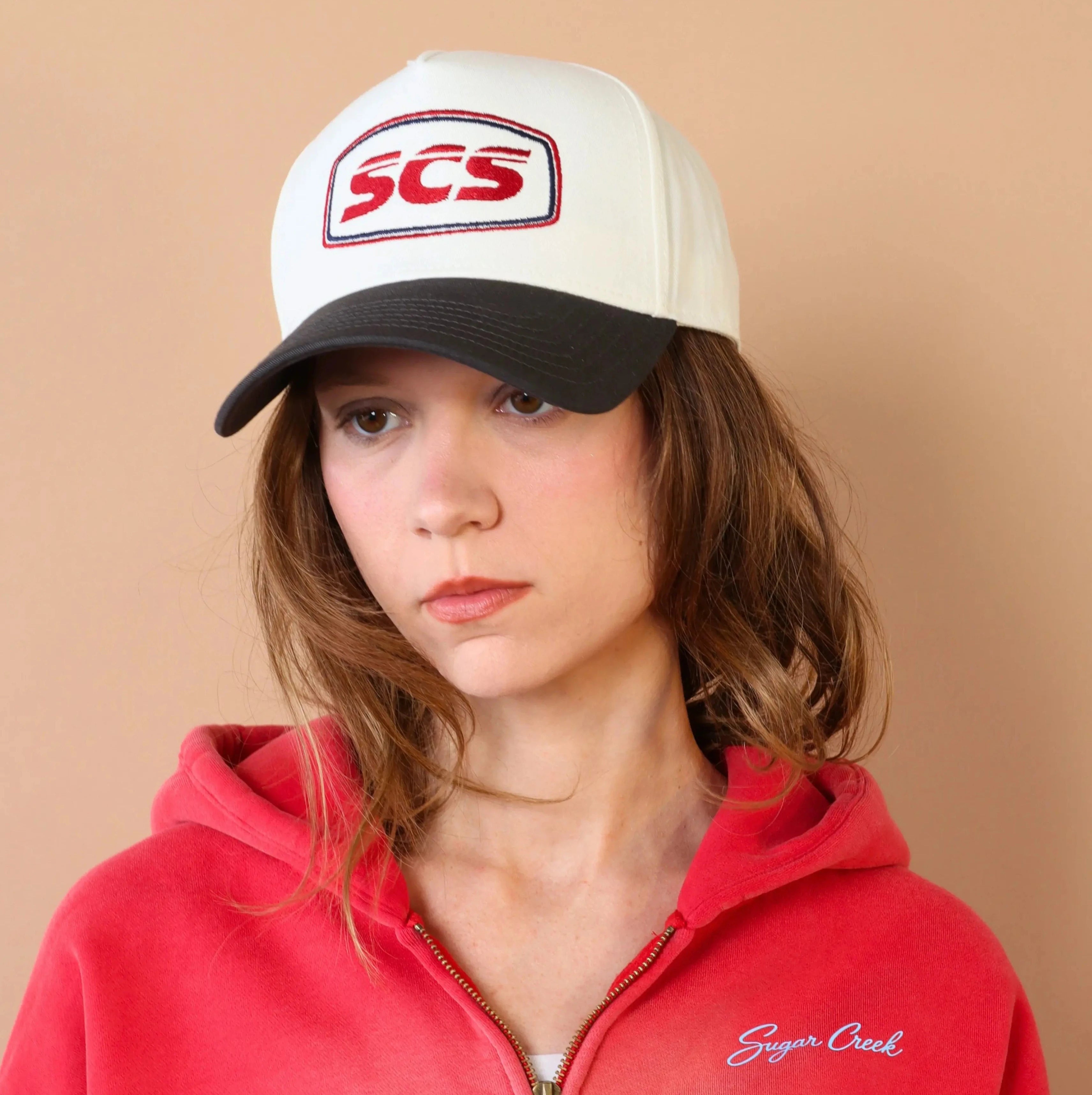 Person wearing a red hoodie and white cap with 'SCS' logo on a beige background