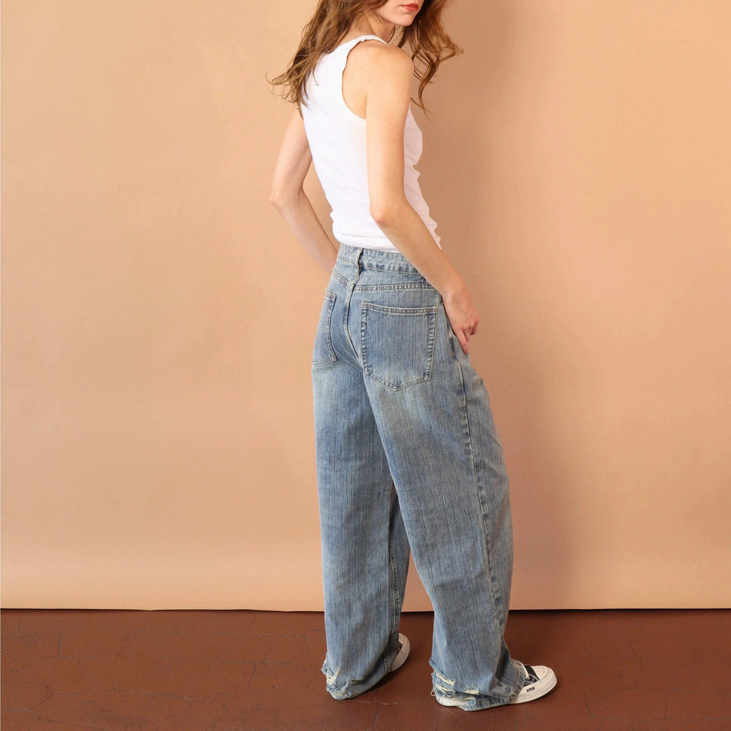 Woman wearing a white tank top and blue jeans against a beige wall.