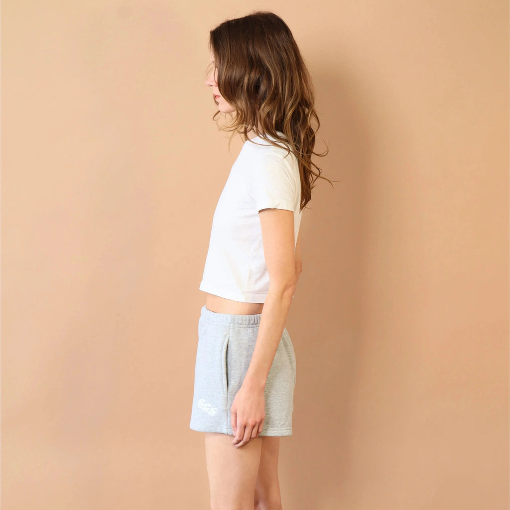 Woman wearing a white top and light blue shorts against a beige wall.