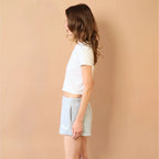 Woman wearing a white top and light blue shorts against a beige wall.