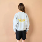 Person wearing a light gray sweatshirt with a logo on the back against a beige wall.
