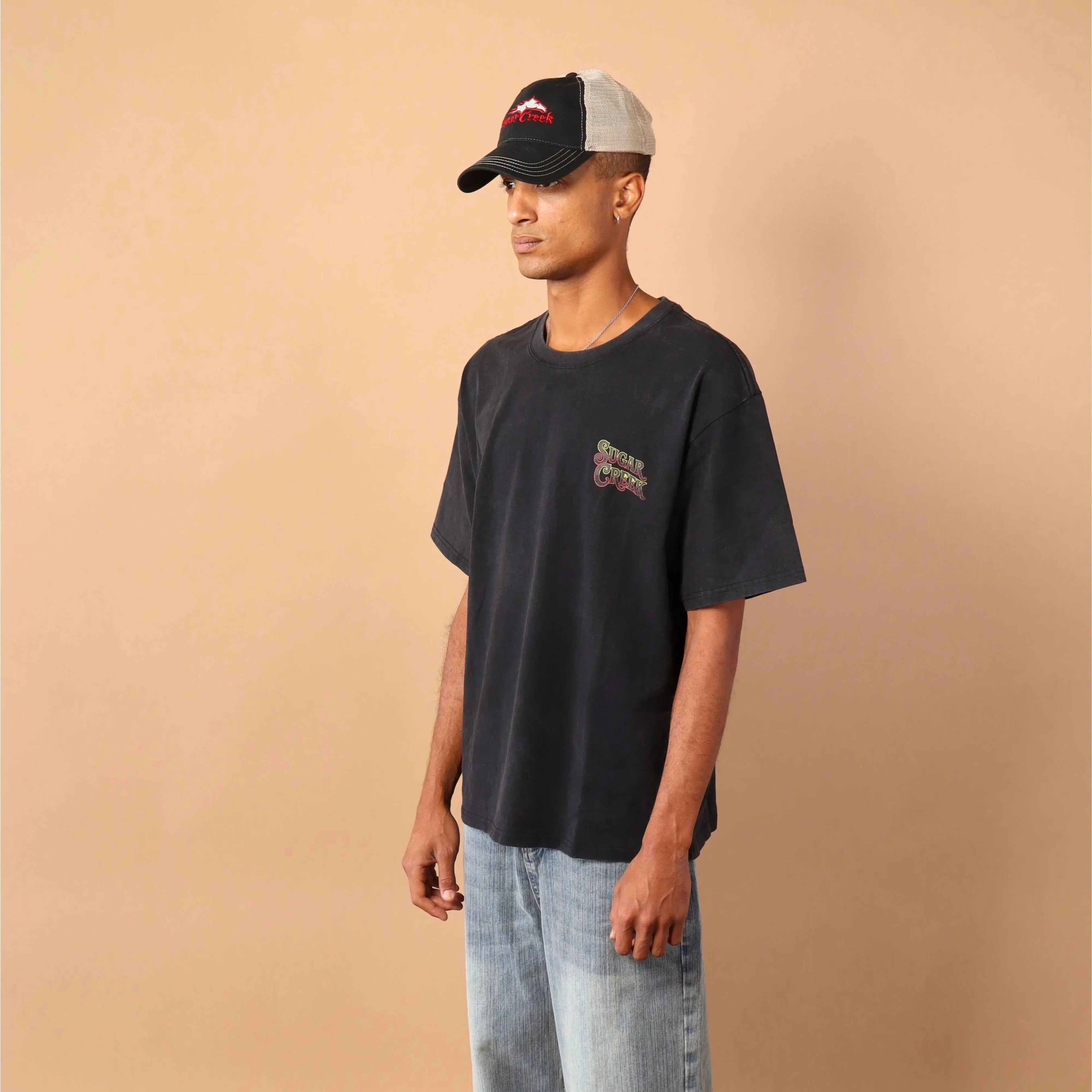 Man wearing a black t-shirt with a logo and light blue jeans against a beige wall.