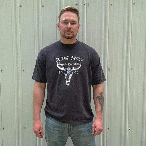 Man wearing a black t-shirt with a graphic design standing against a metallic wall.