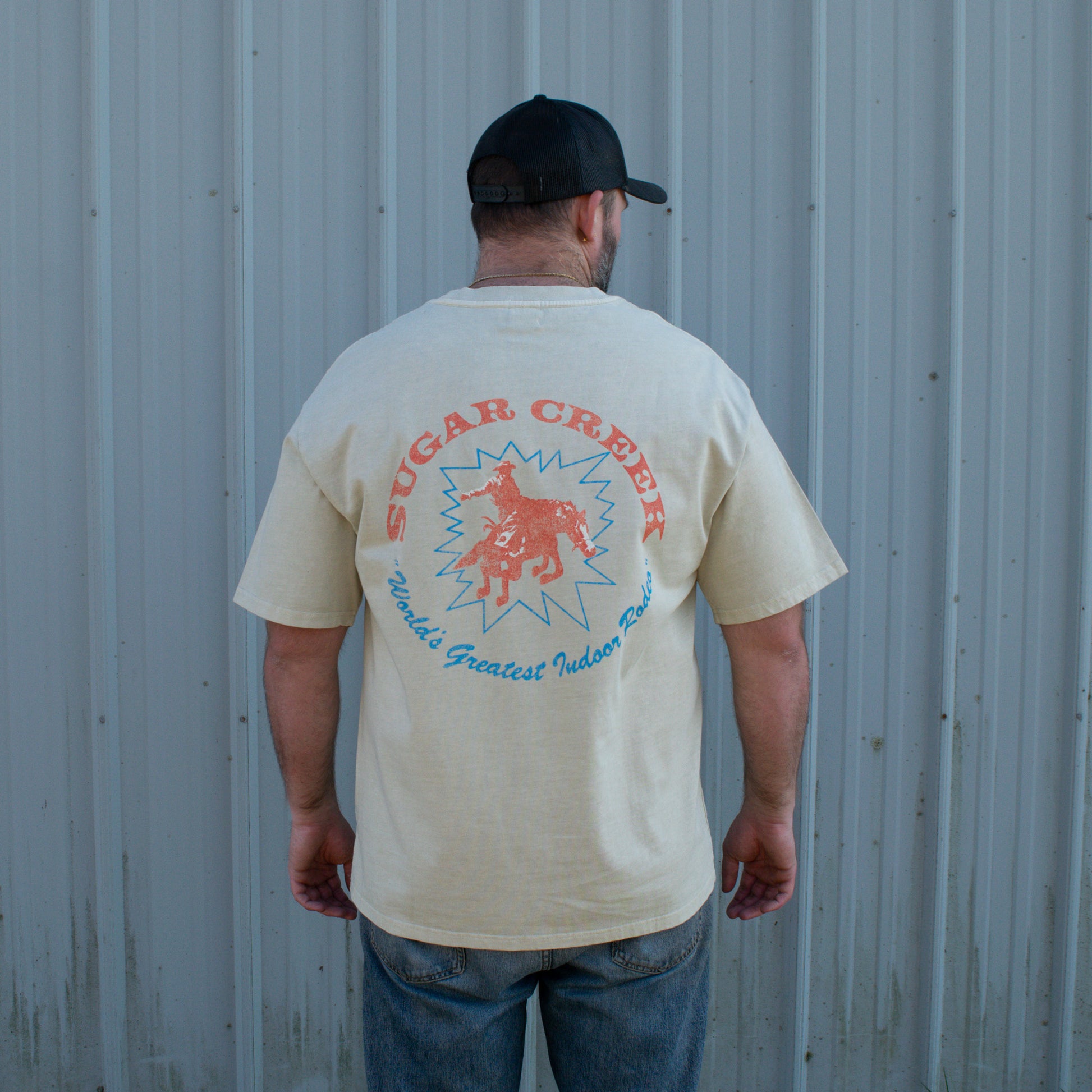 Man wearing a beige t-shirt with 'Sugar Creek' graphic design against a gray wall.