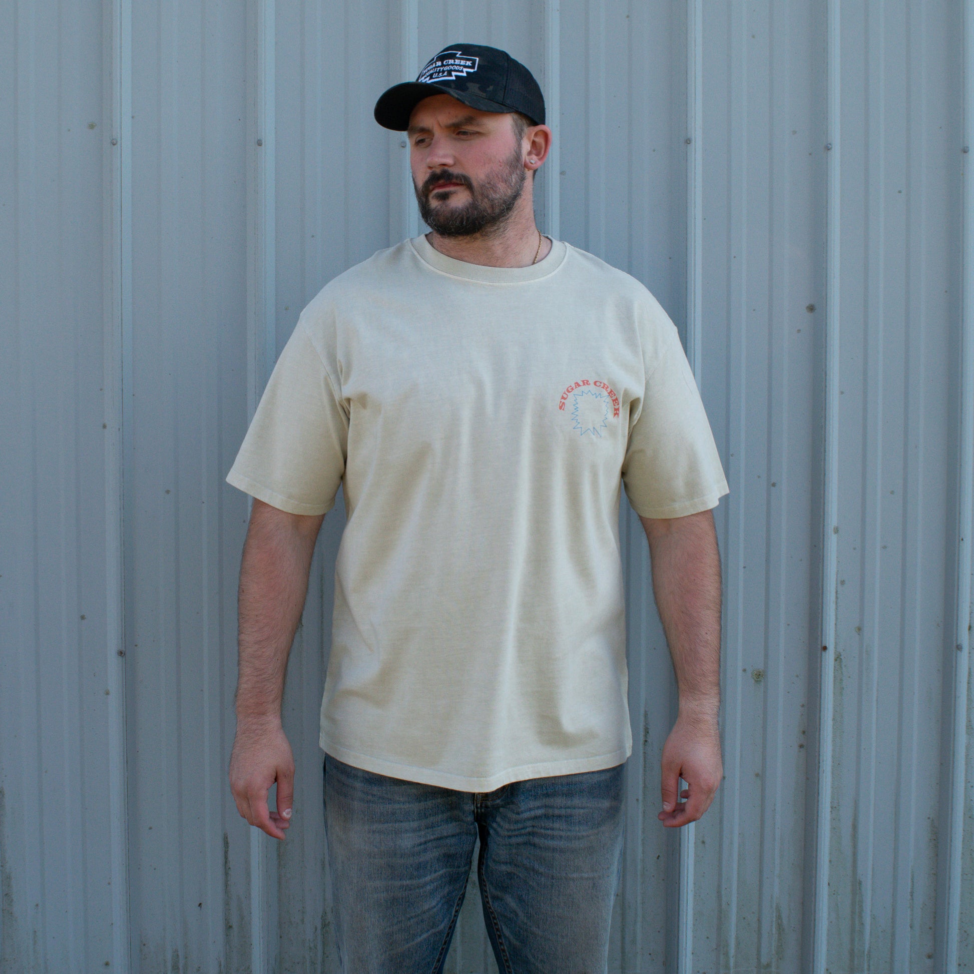 Man wearing a beige t-shirt with a logo and a black cap, standing against a corrugated metal wall.