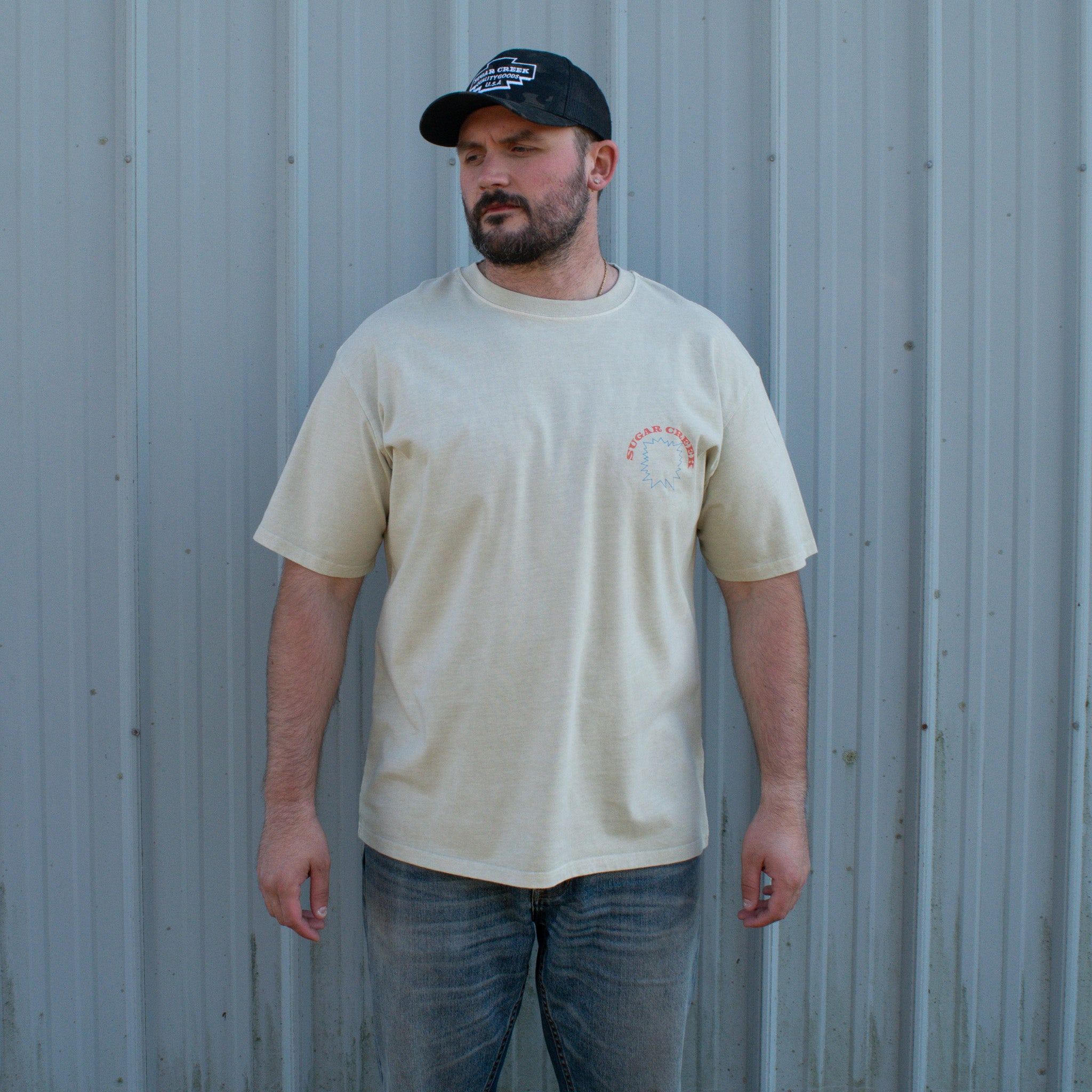 Man wearing a beige t-shirt with a logo and a black cap, standing against a corrugated metal wall.