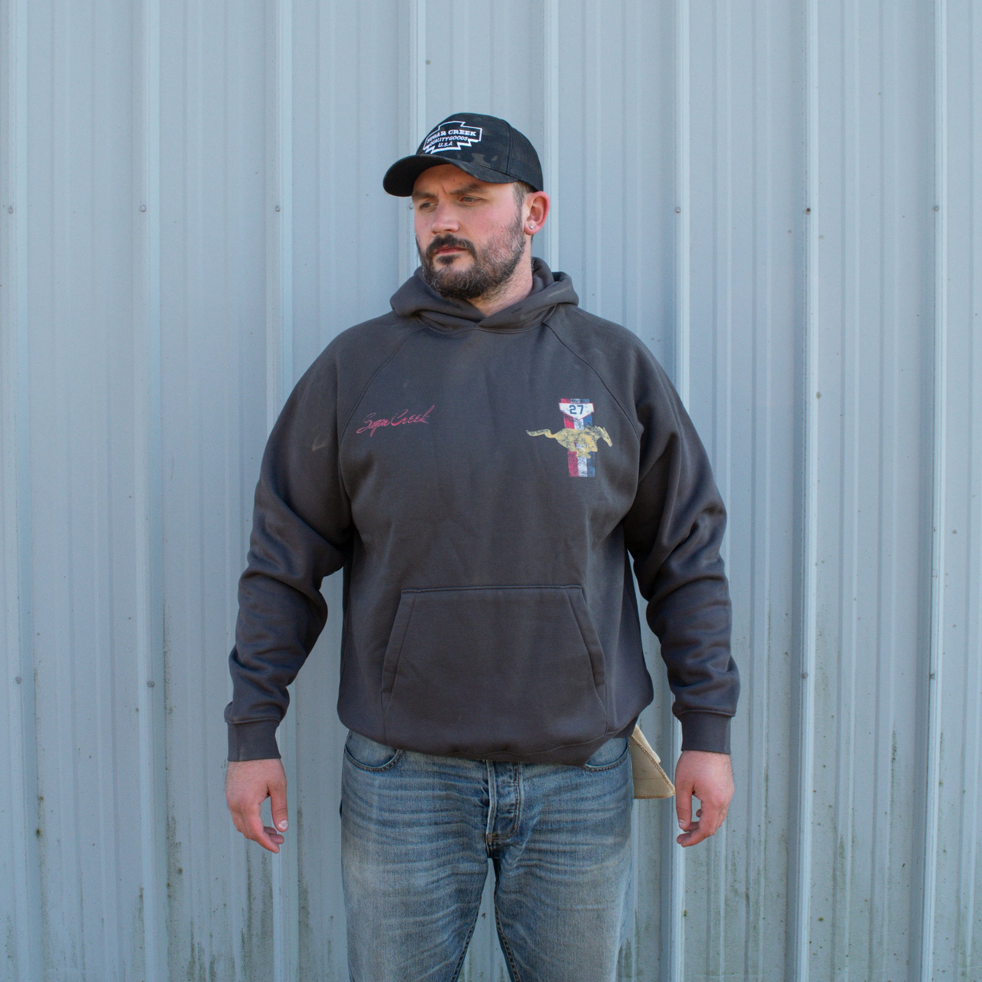 Man wearing a dark hoodie with a logo and a cap, standing against a corrugated metal wall.
