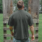 Person wearing a green t-shirt and light blue jeans standing between two wooden pallets in an outdoor setting.
