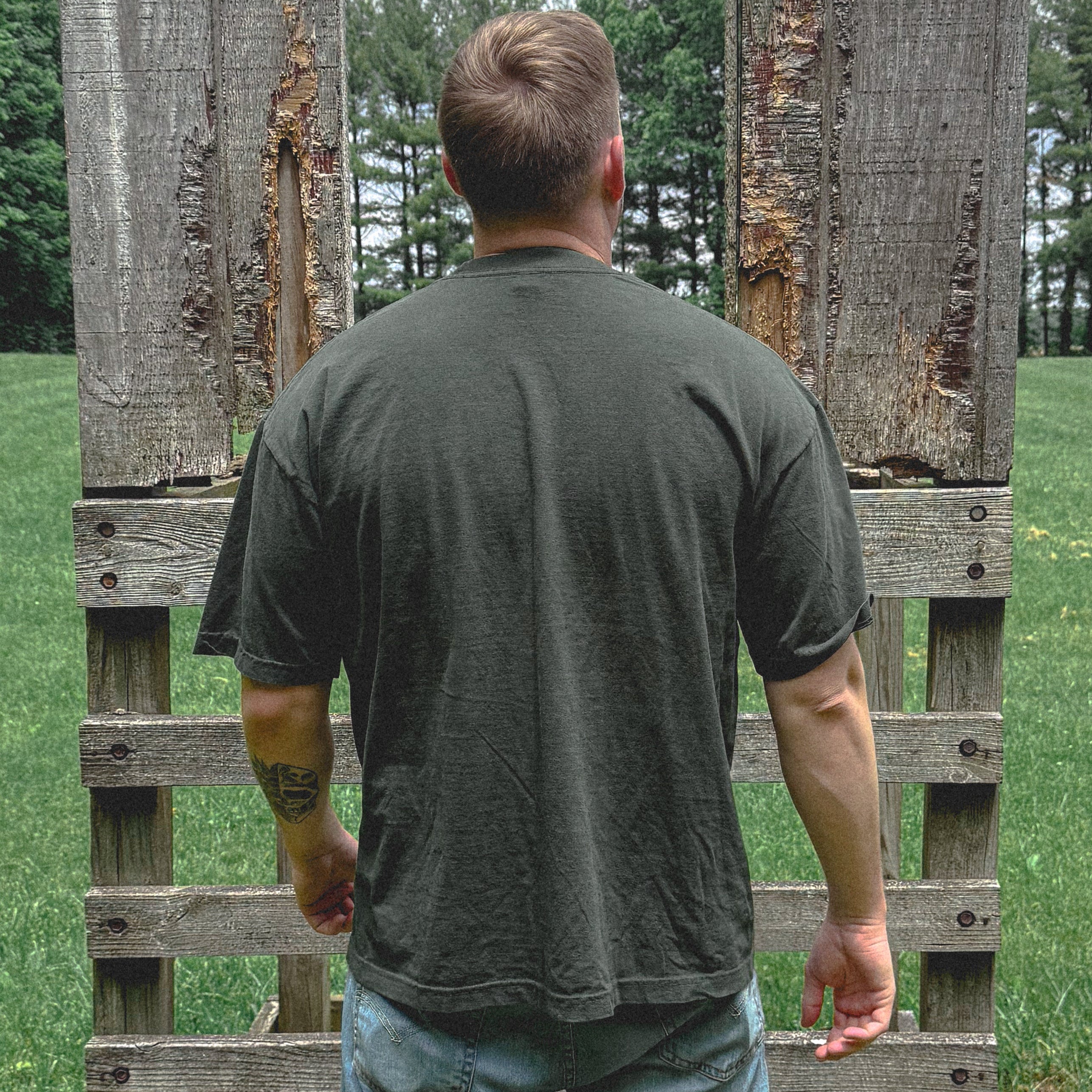 Person wearing a green t-shirt and light blue jeans standing between two wooden pallets in an outdoor setting.