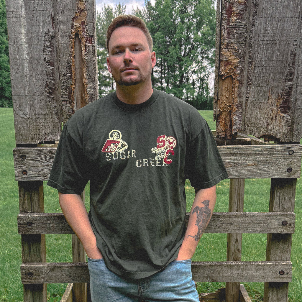 Man wearing a dark green t-shirt with text and graphics, standing in front of wooden pallets.